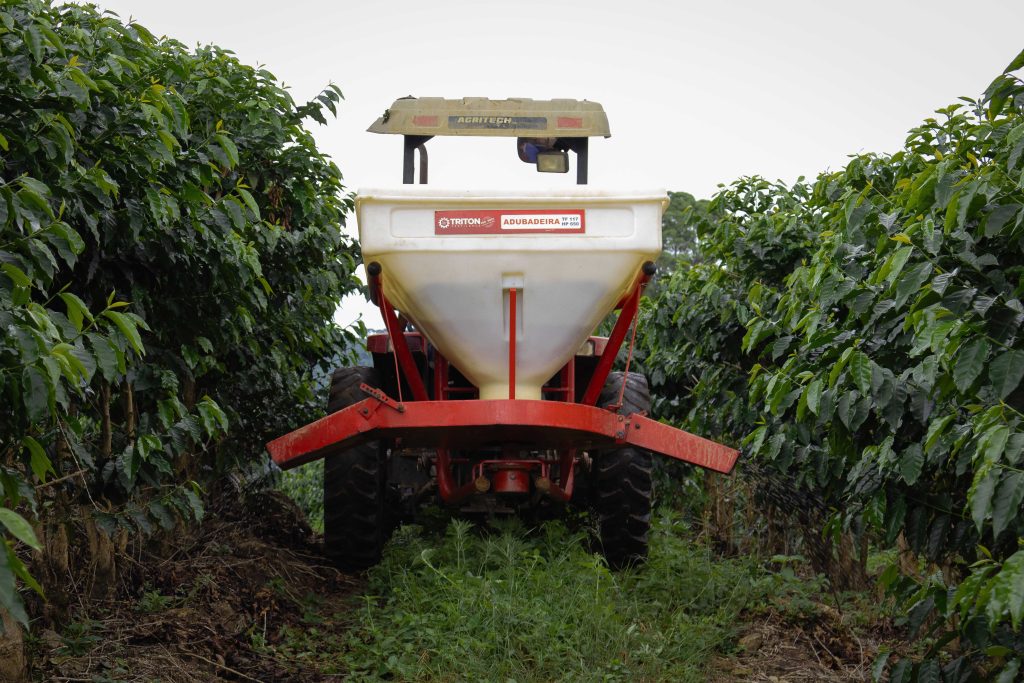Agricultural tractor equipped with a fertilizer spreader moving between rows of tall, green coffee plants in a plantation.