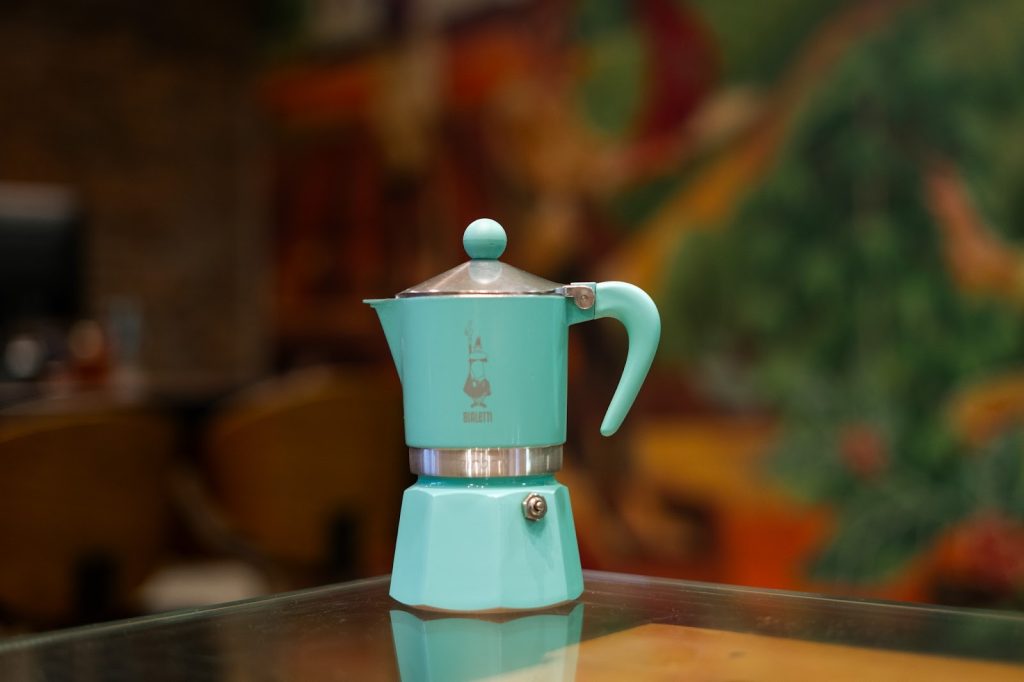 Uma cafeteira Moka Express Bialetti verde-menta de fogão sobre uma mesa de vidro em um ambiente de café desfocado.