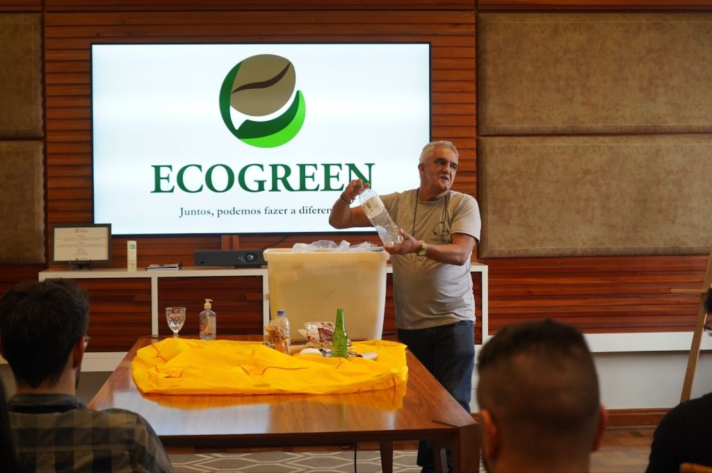 An environmental education presentation for the EcoGreen project at Bourbon Specialty Coffees. A speaker demonstrates recycling practices with plastic bottles to an audience, with a large screen displaying the EcoGreen logo and the slogan 'Together, we can make a difference