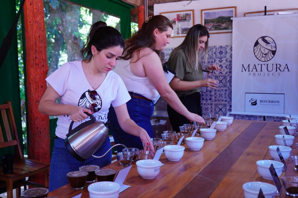 Profissionais de café mulheres participam de uma sessão de cupping e treinamento de café especial, derramando água em xícaras sobre uma mesa longa. Ao fundo, um banner exibe os logotipos 'Projeto Matura' e 'Bourbon', ilustrando empoderamento e educação de qualidade